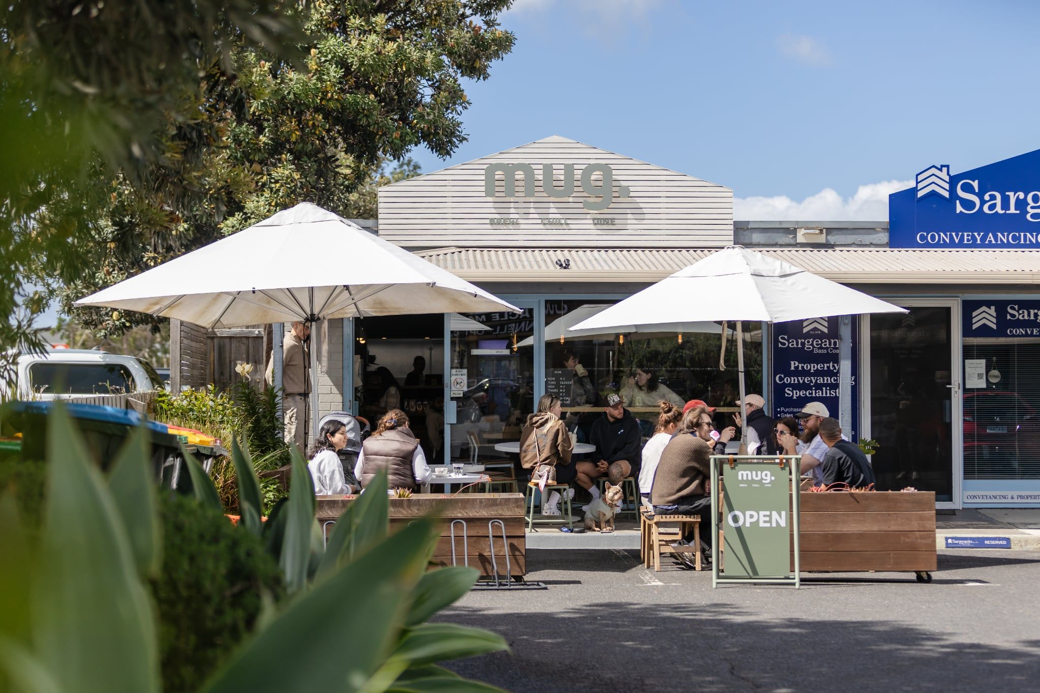 mug. café exterior with customers dining under white umbrellas on a sunny day in Cowes