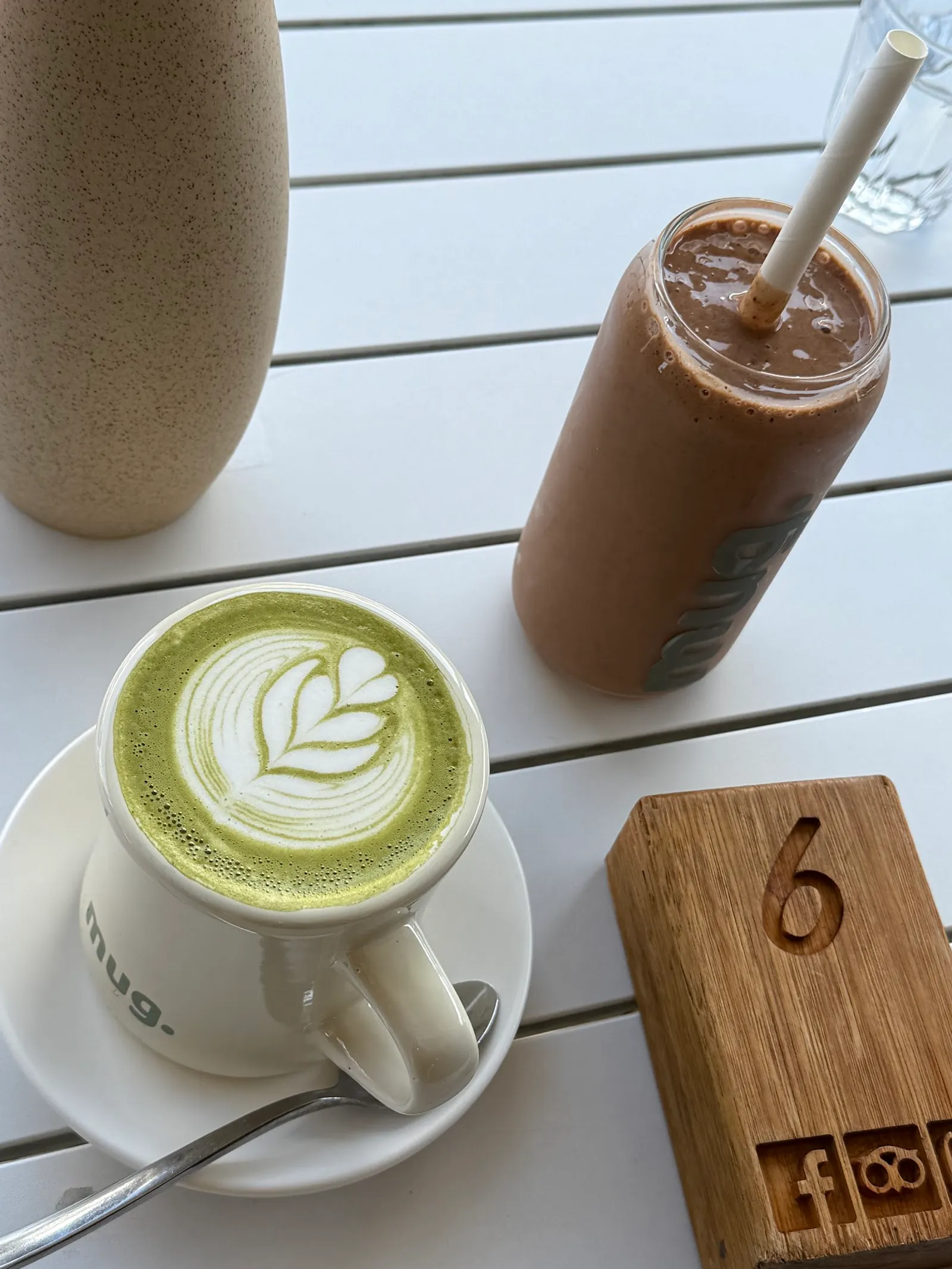 Matcha latte with latte art in a mug. branded cup next to an iced chocolate drink on a white outdoor table