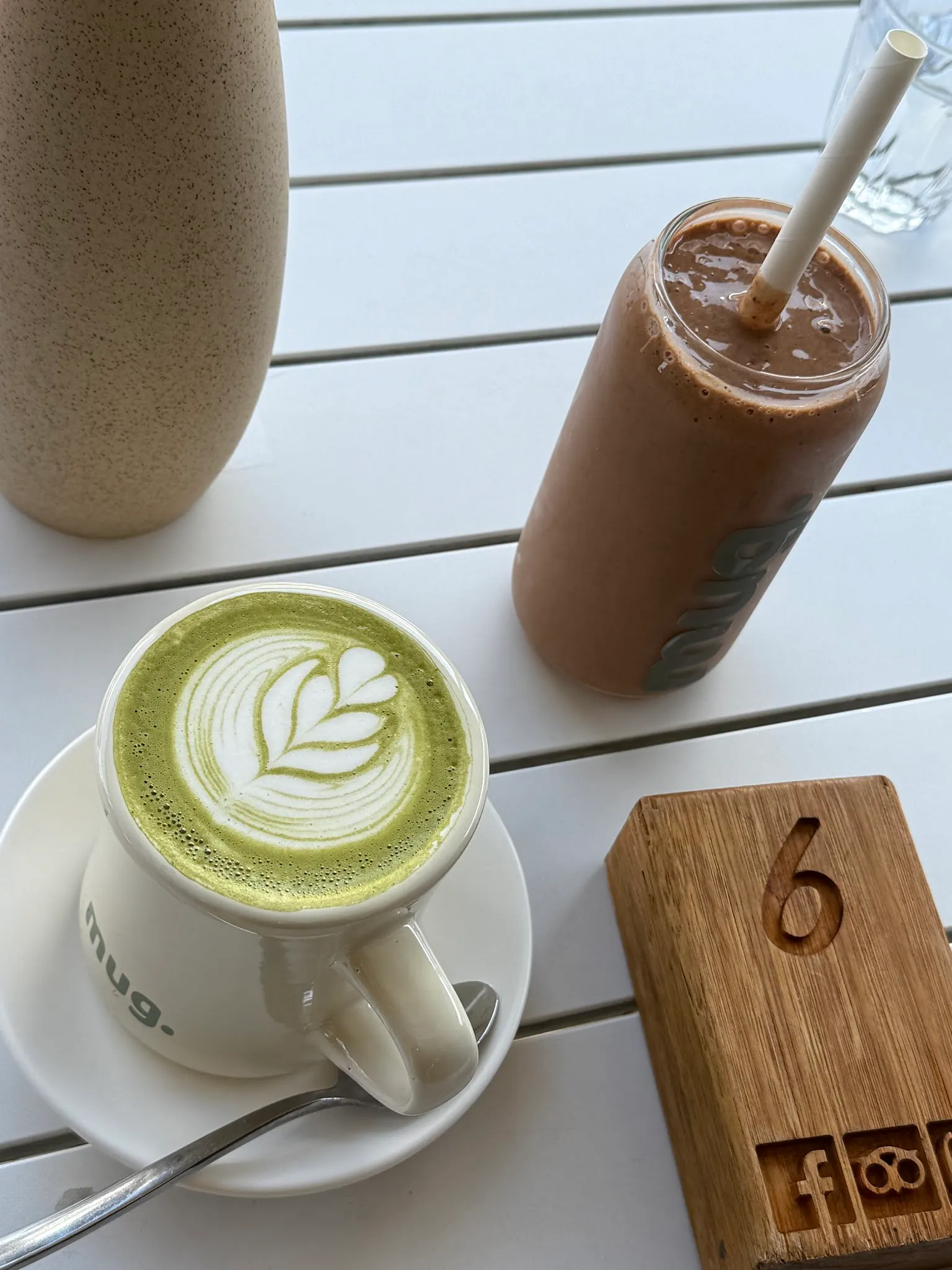 Matcha latte with latte art in a mug. branded cup next to an iced chocolate drink on a white outdoor table