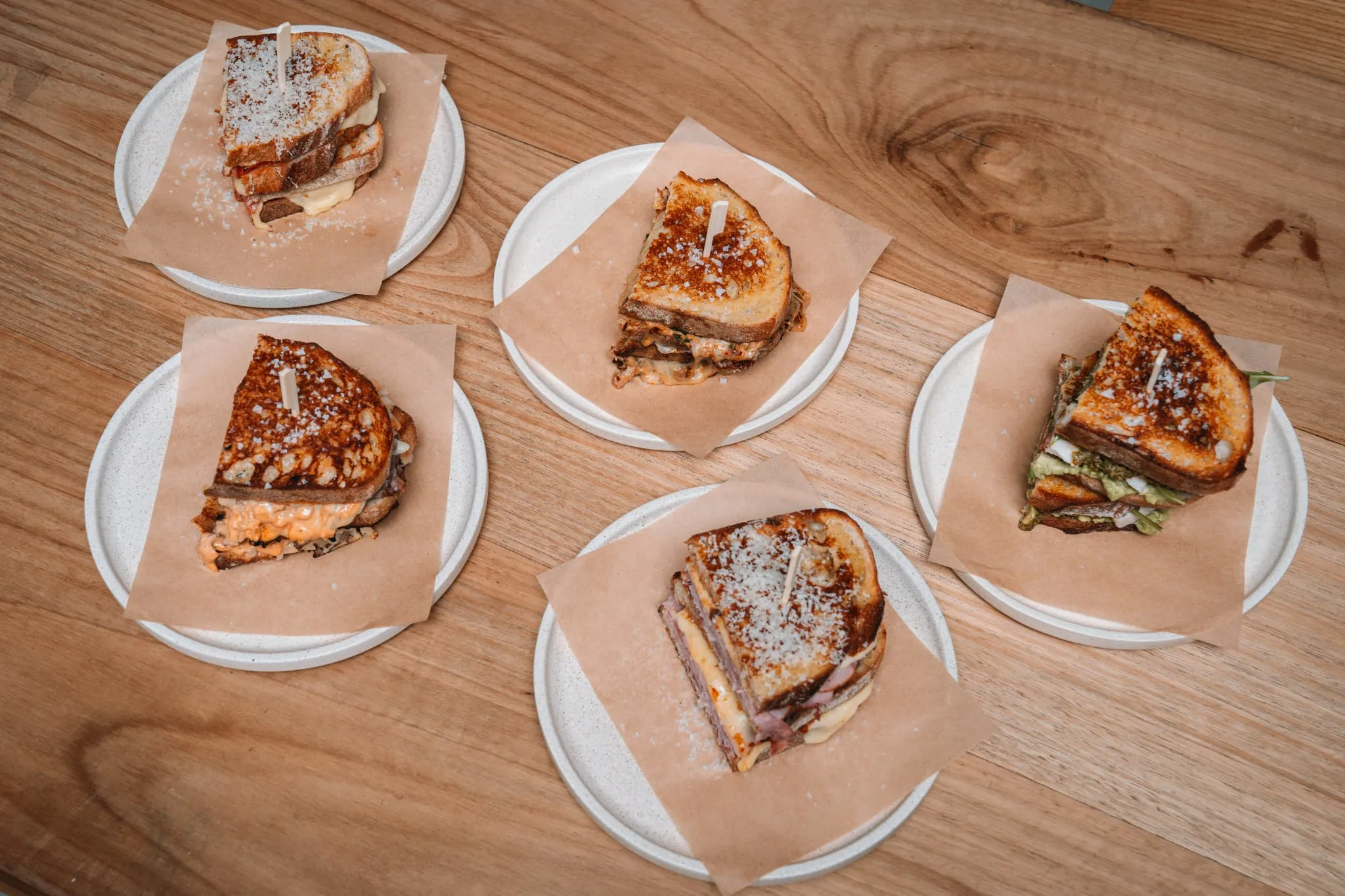 Five different toastie varieties arranged on plates with parchment wrappers, overhead view on timber table