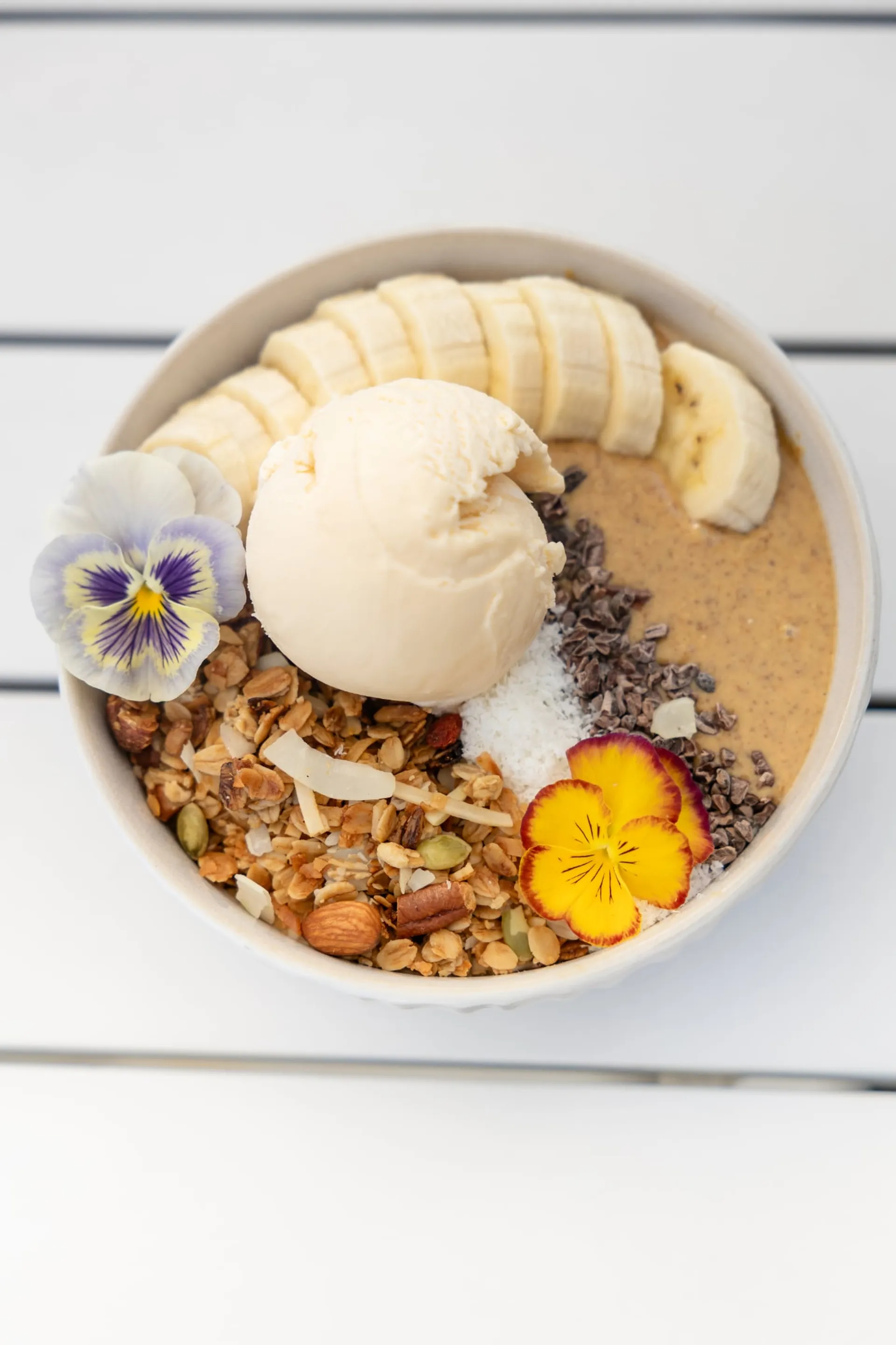 Smoothie bowl with banana, granola, coconut, cacao nibs, ice cream and edible flowers
