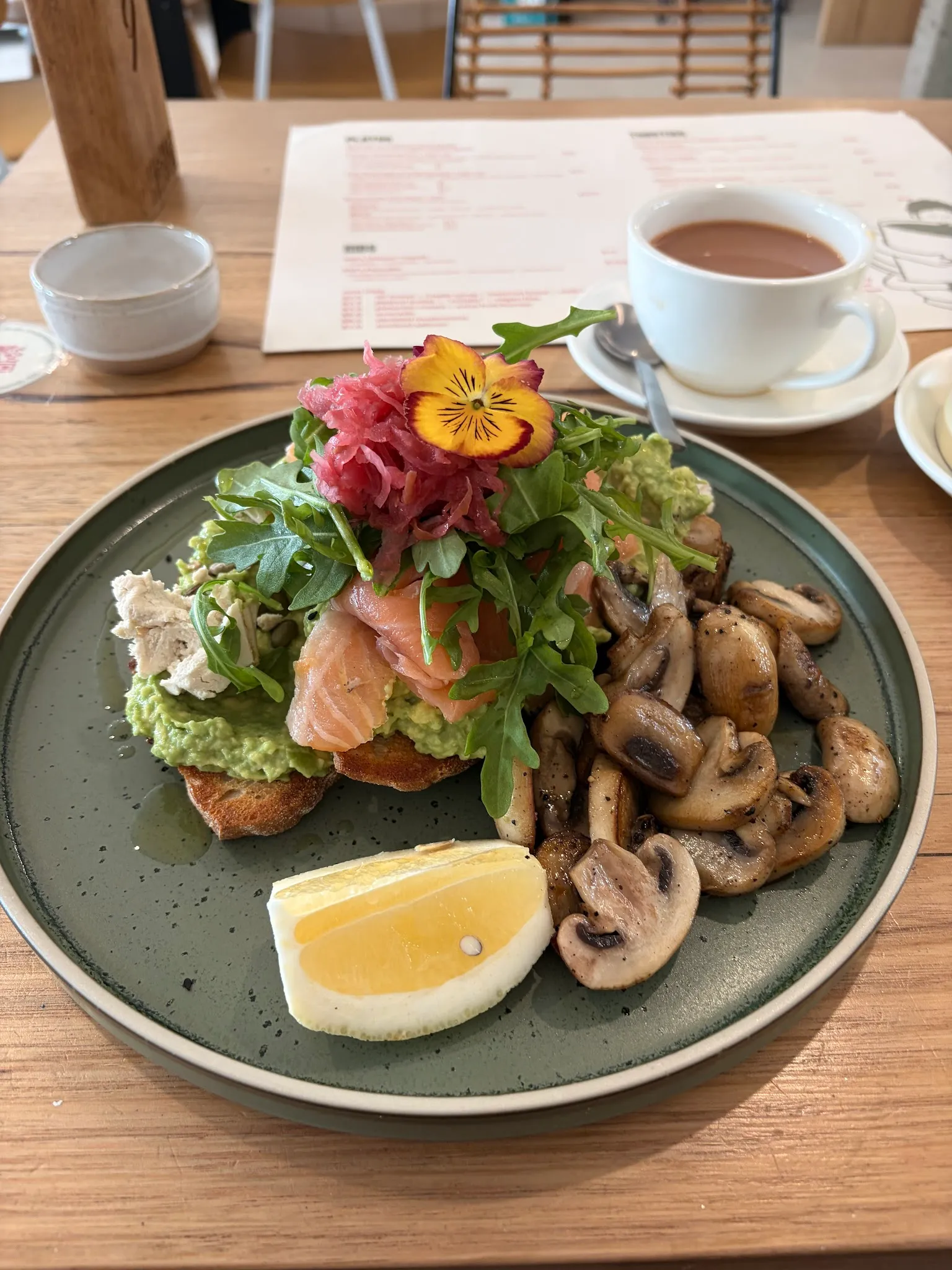 Smashed avocado with salmon, mushrooms and herbs on a sage plate with hot chocolate in the background
