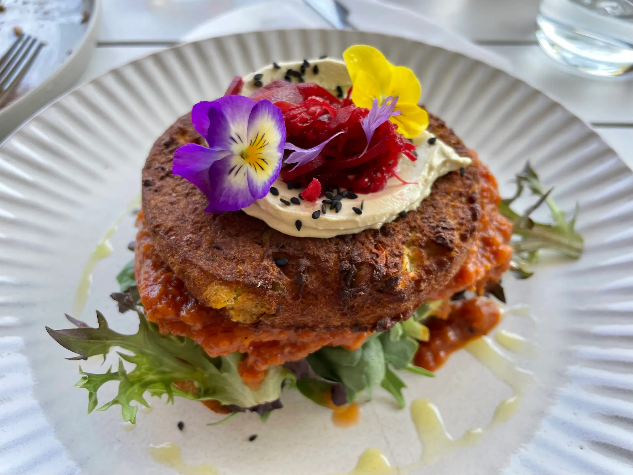 Vegan fritter stack with whipped cream, beetroot relish and edible flowers on a white ribbed plate