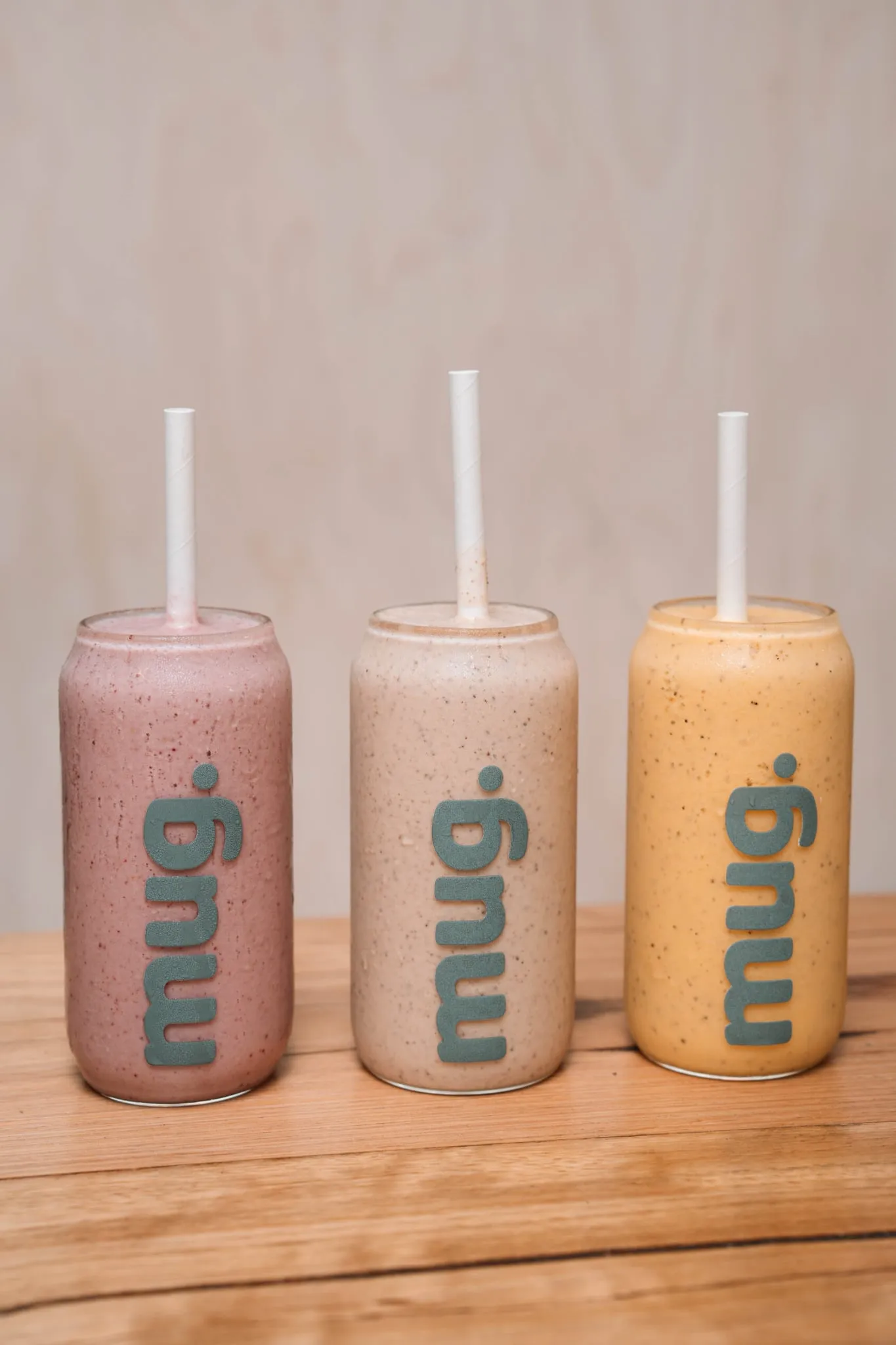 Three mug. branded smoothie glasses in pink, cream and golden yellow lined up on a timber surface