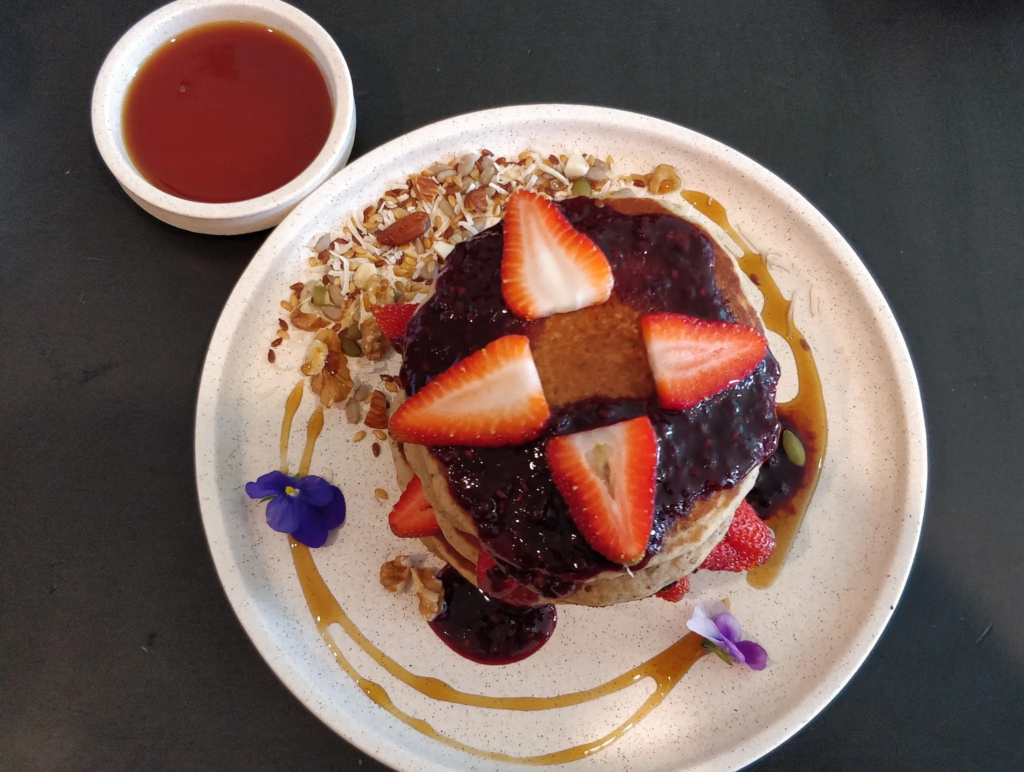 Pancakes with strawberry, blueberry compote, granola and edible flowers on a white plate with syrup ramekin