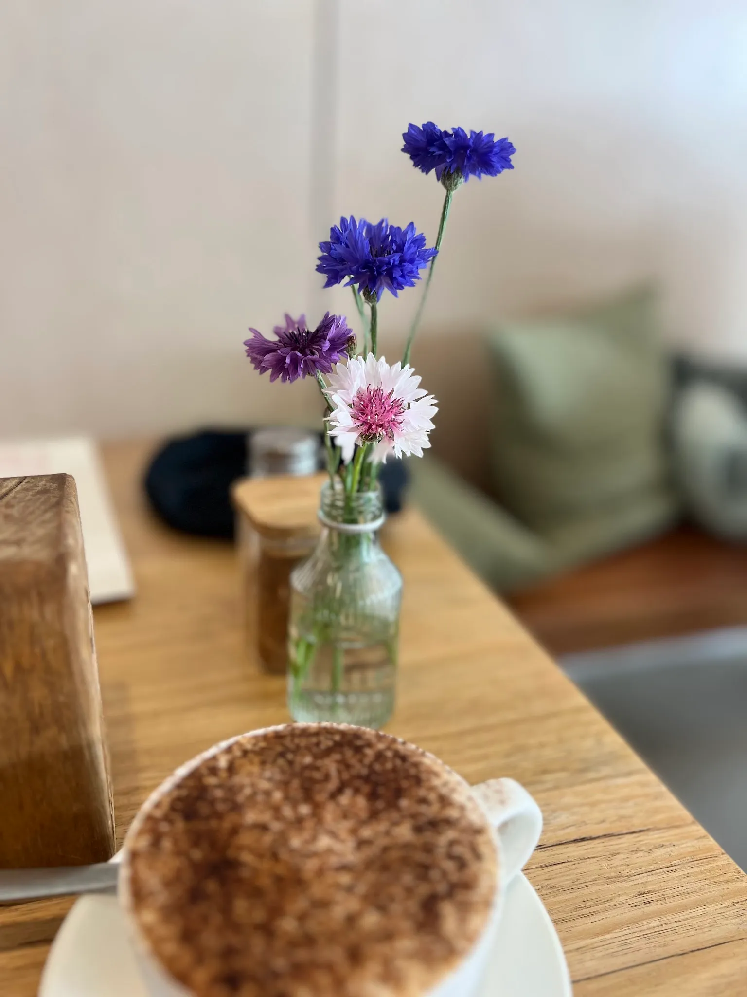Blue and purple wildflowers in a glass bottle vase on a timber café table with a coffee cup in soft focus foreground